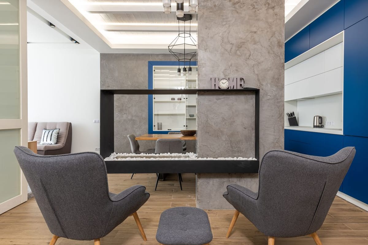 Two grey wingback chairs face a concrete room divider with a pass-through opening; blue cabinetry and a dining table are visible beyond.