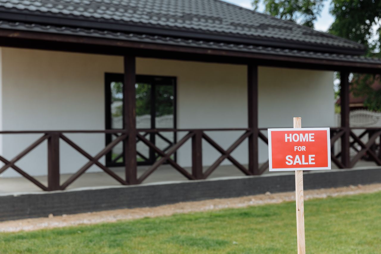 A red Home For Sale sign on a wooden stake in front of a white house with a covered porch and dark wood railings.
