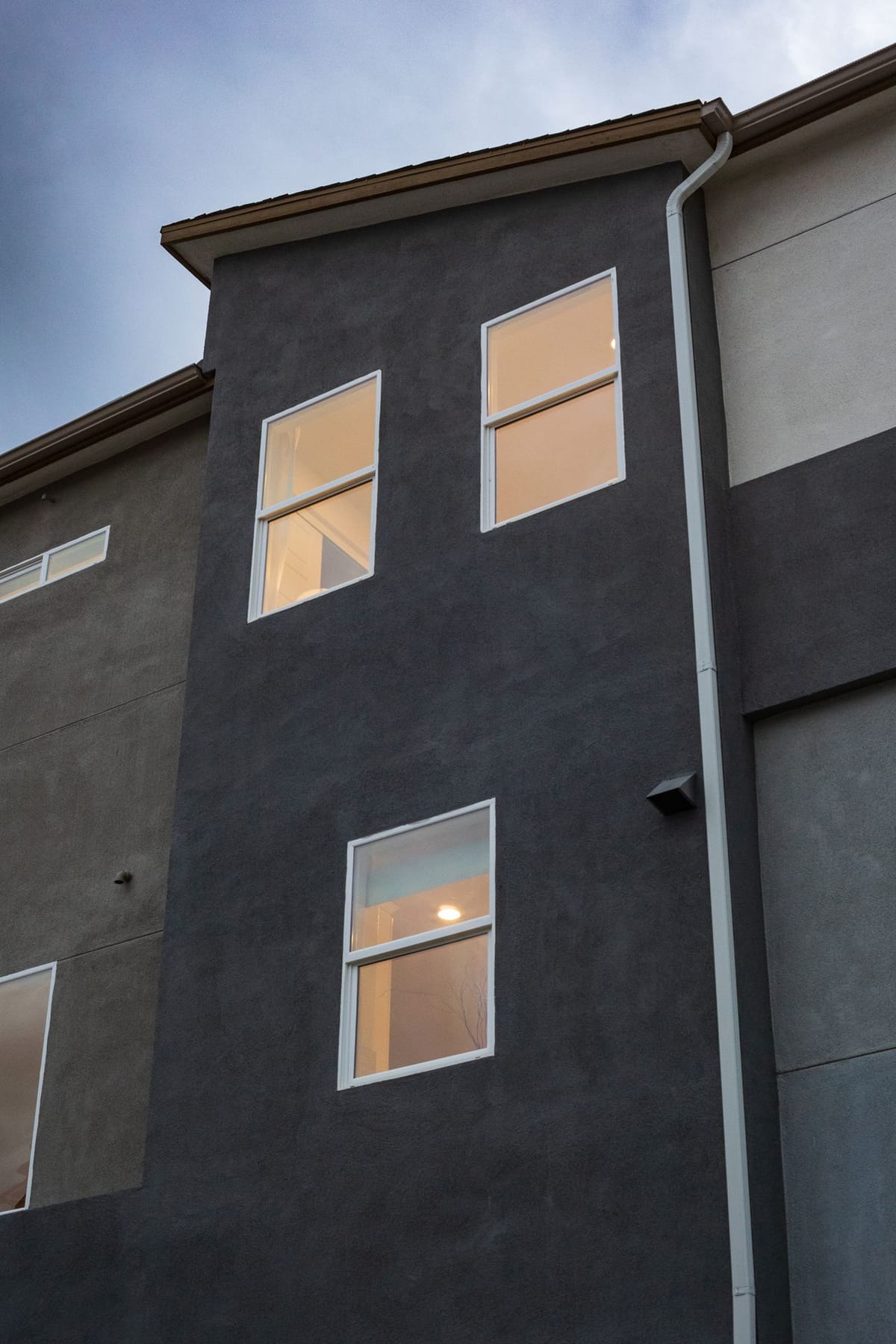Low-angle view of a dark grey stucco building exterior at dusk with lit windows and a white downspout.