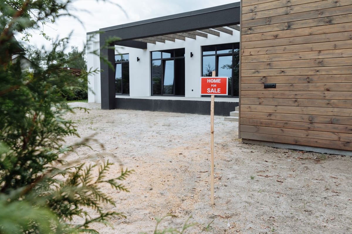 A red Home For Sale sign on a stake in front of a modern house with wood cladding, dark trim, and a gravel yard.